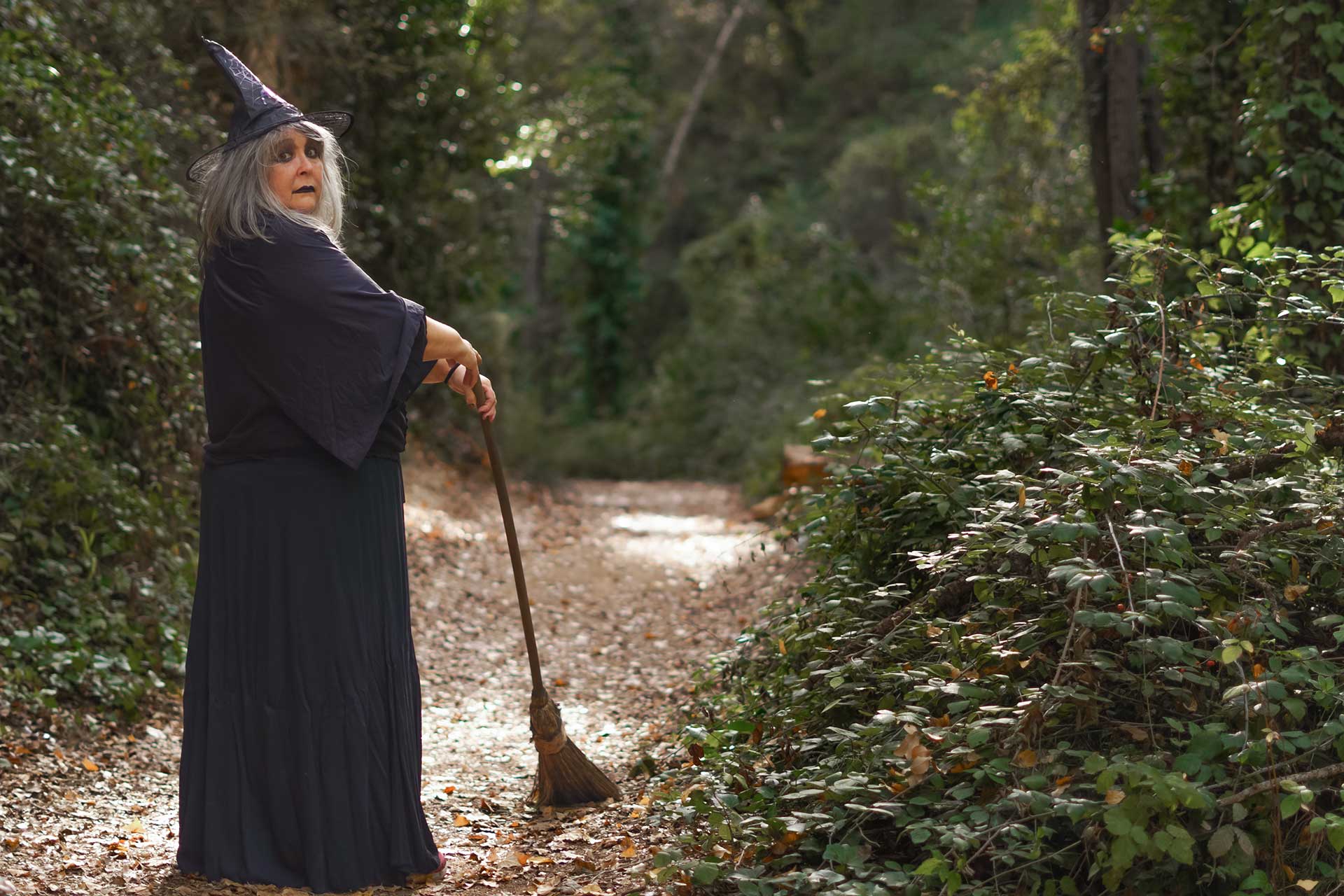 An older woman in a witch's costume stands on a path in the woods holding a broomstick.