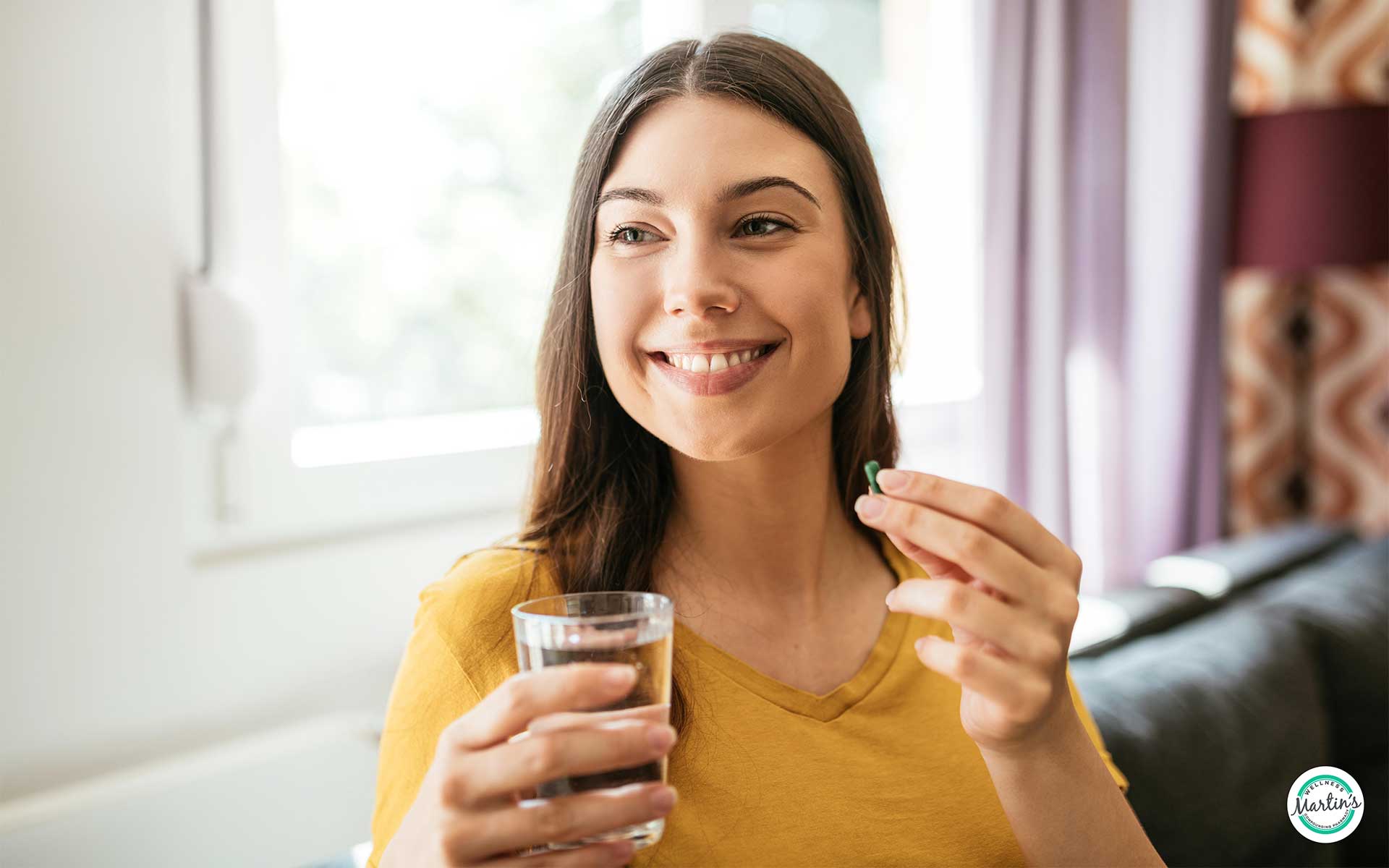 A smiling brunette woman sitting in a sunny room holding up an NAD+ precursor supplement capsule and a glass of water