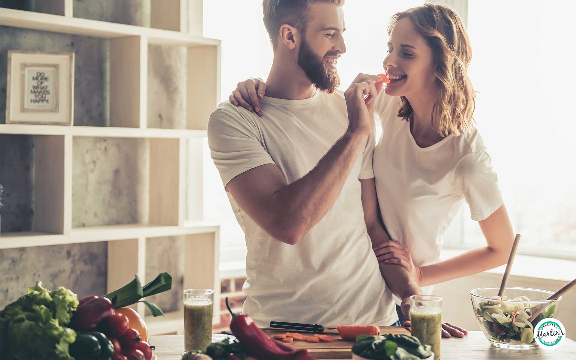 A couple in white shirts, smiling in a kitchen. The man feeds the woman a slice of bell pepper