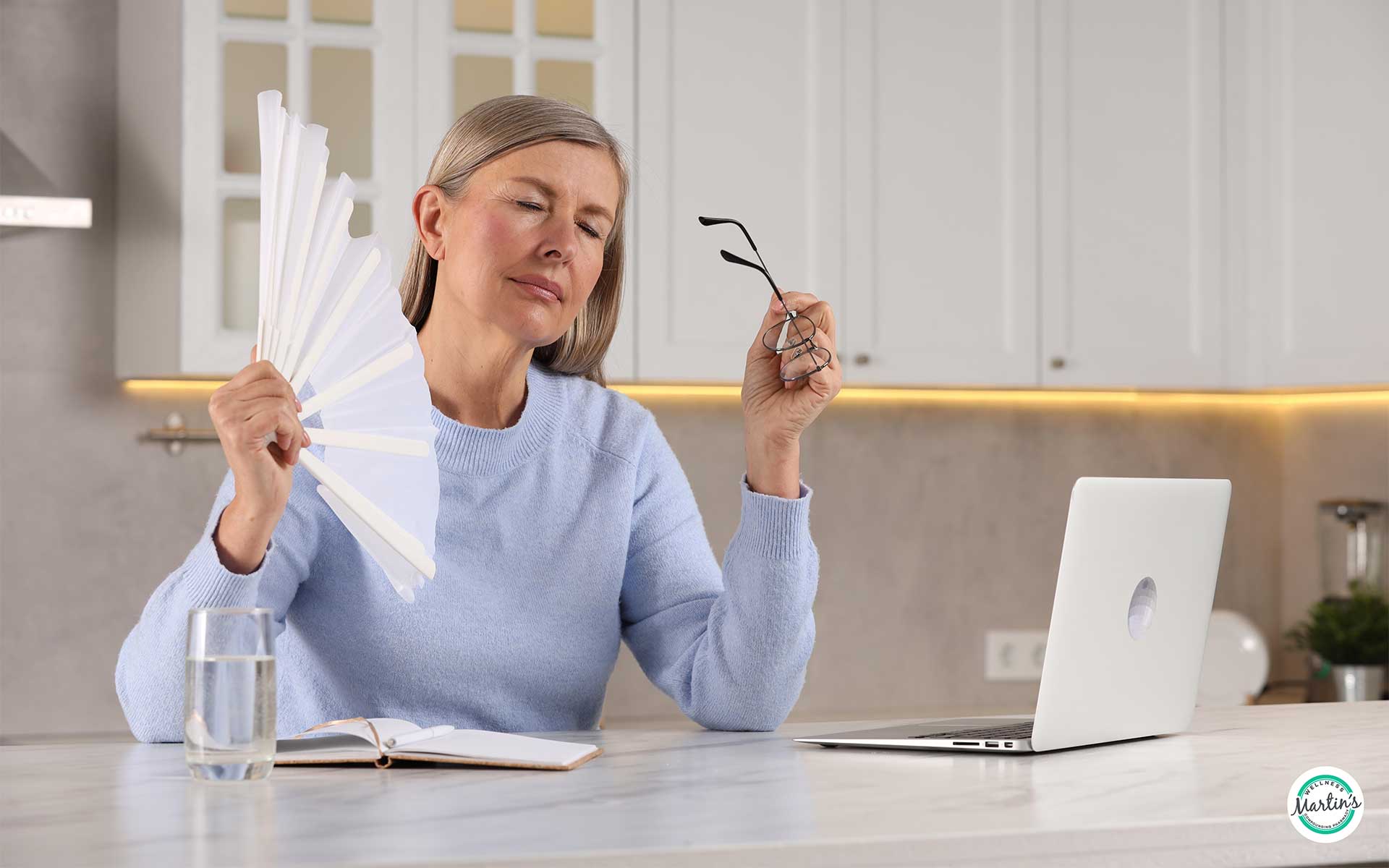 Middle-aged woman experiencing a hot flash while working at home, using a hand fan for relief