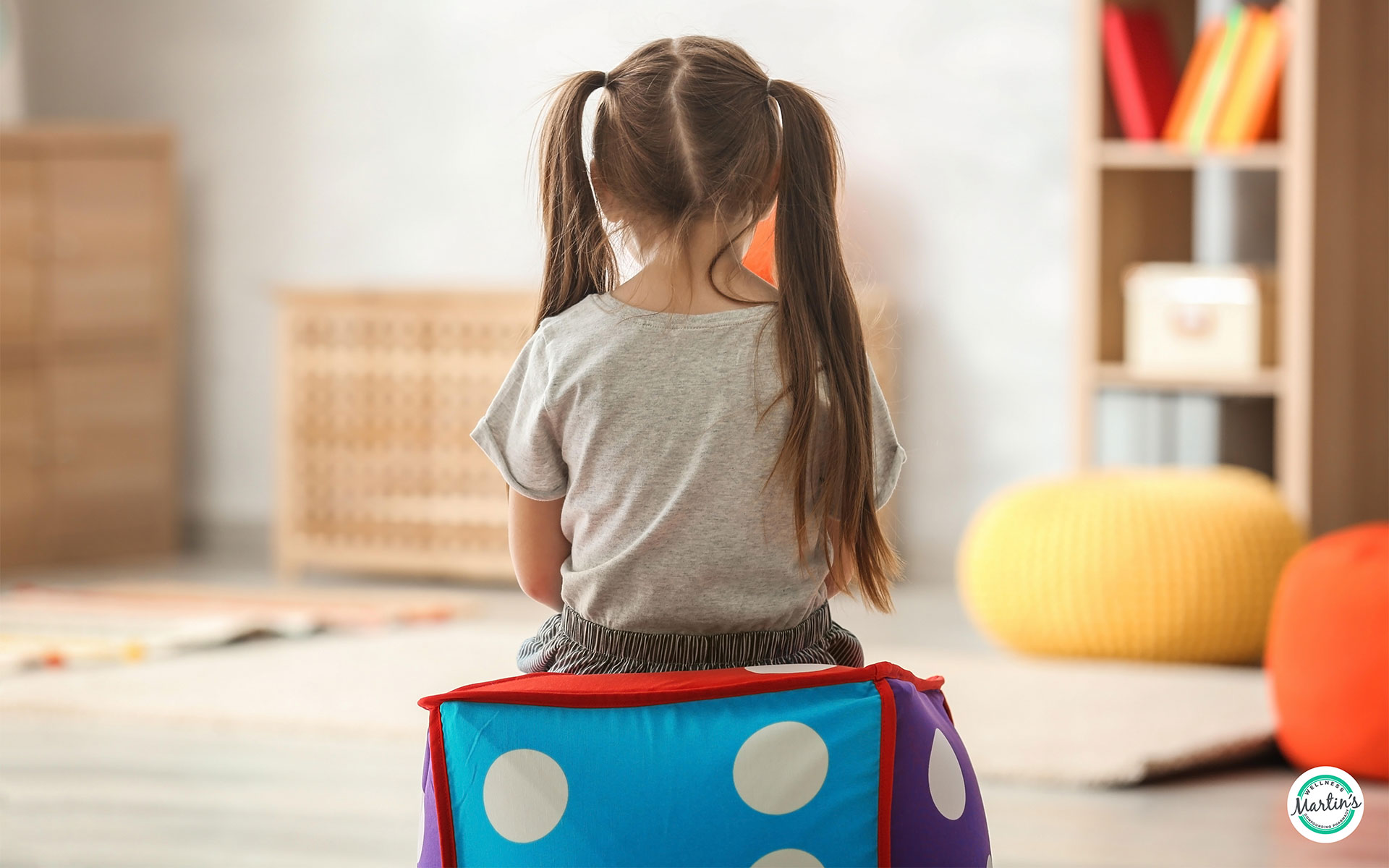 A young girl in pigtails sitting alone on a colorful square cushion with her back facing the camera