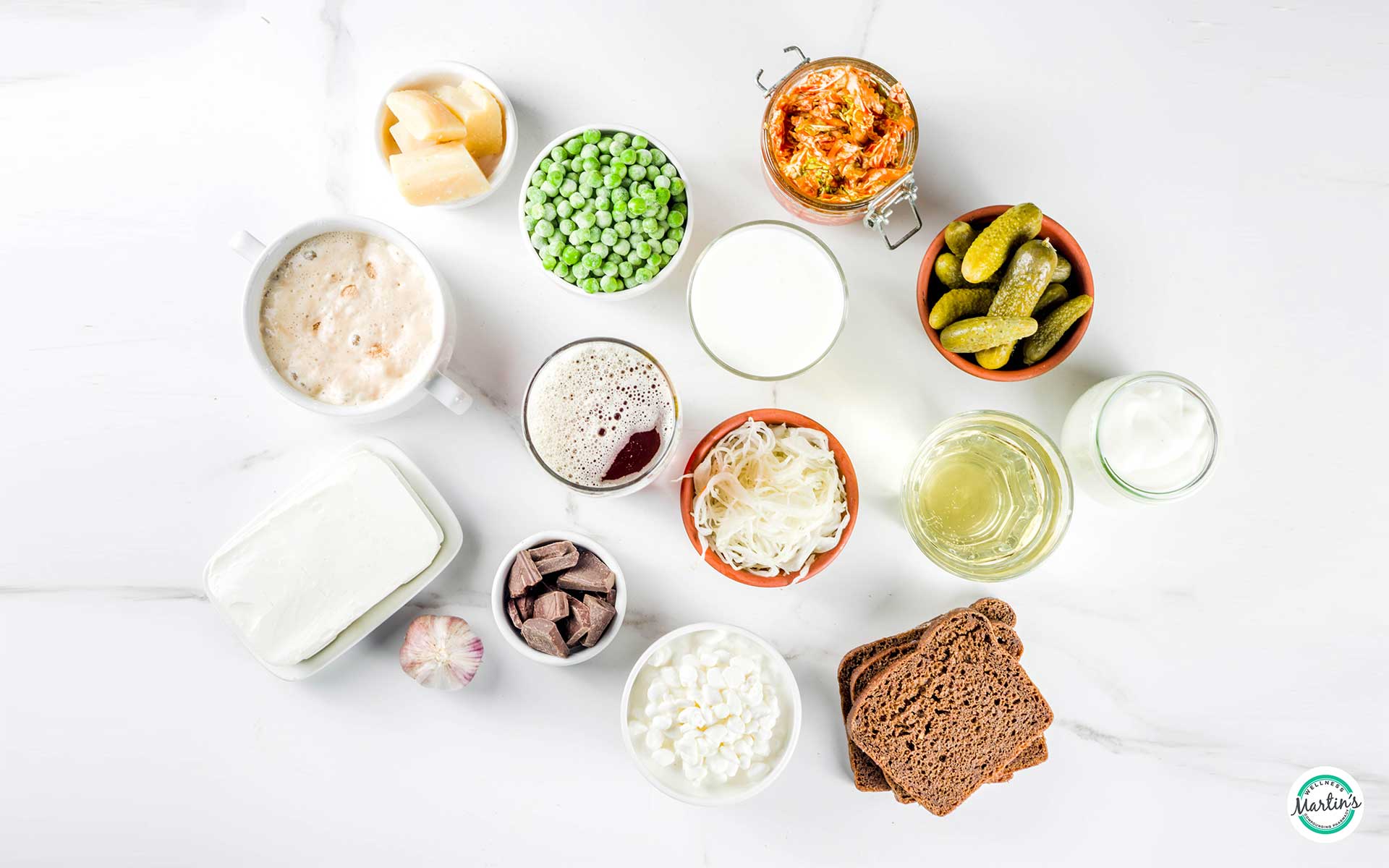 A variety of fermented foods on a white surface, including kimchi, bread, yogurt, sauerkraut, pickles, and chocolate.