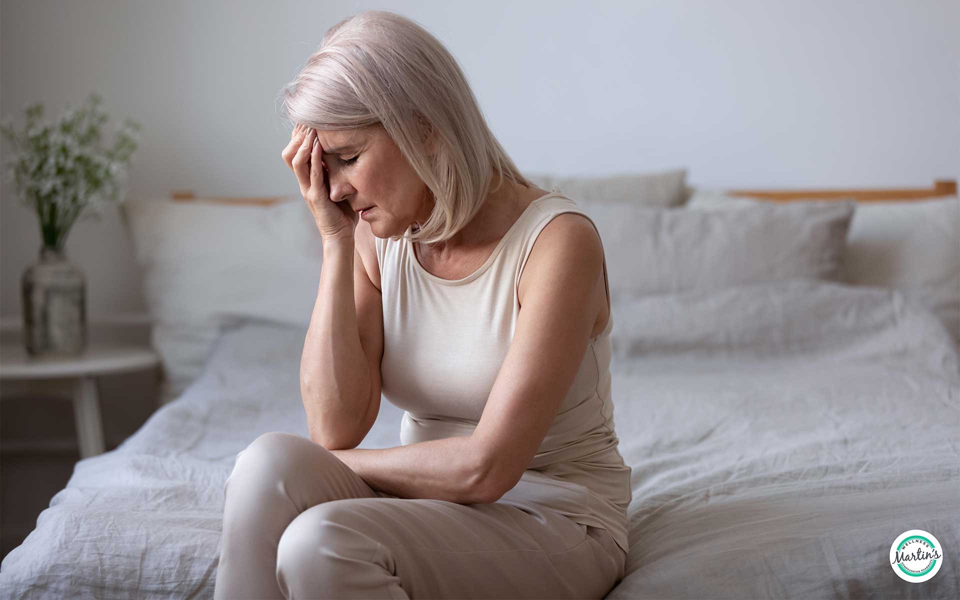 Older woman sitting on bed with hand on forehead, showing signs of fatigue or menopause-related symptoms