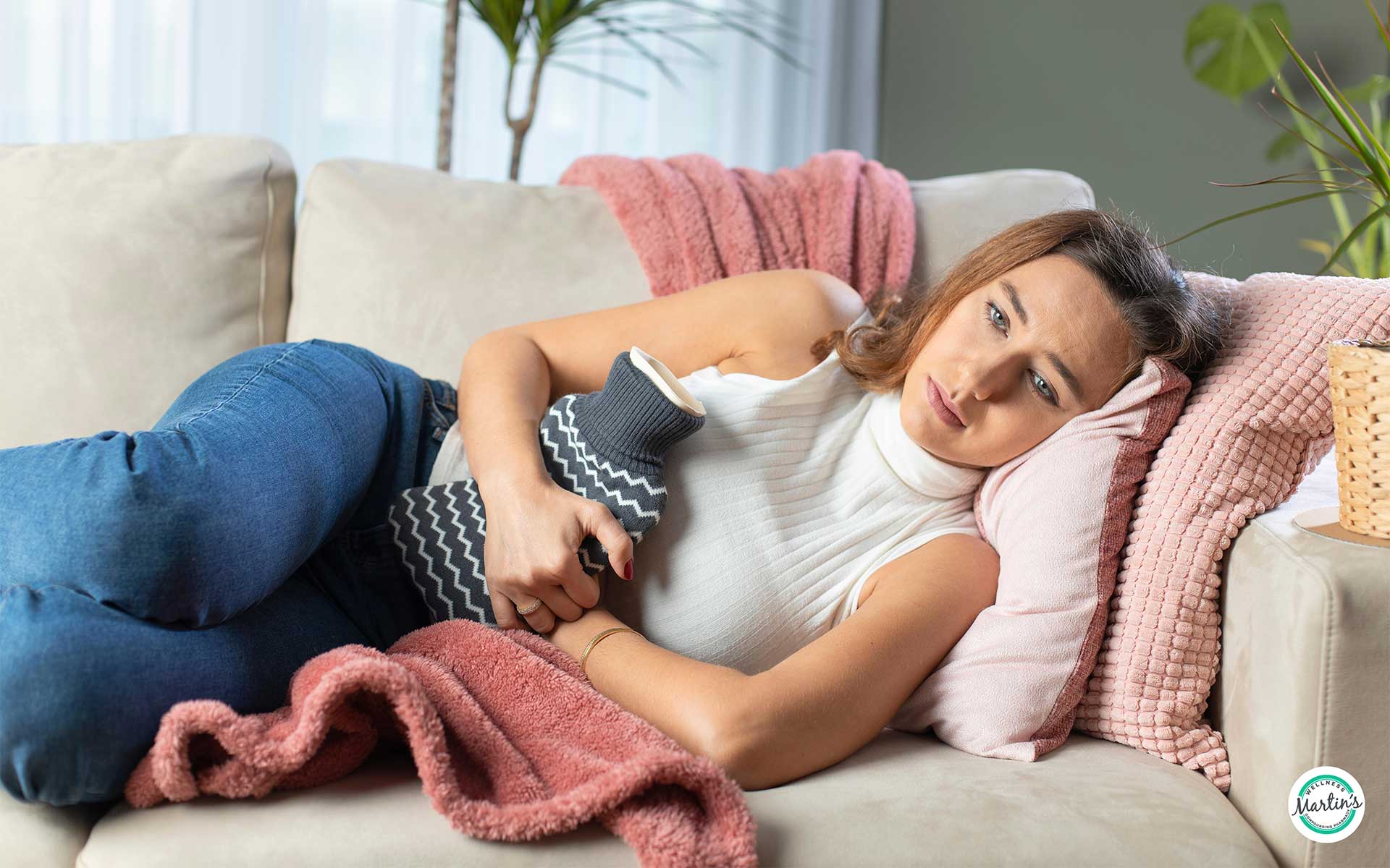 A young woman lies on her side on a couch, knees bent, looking sad as she hugs a heating pad to her belly to ease PMS symptoms
